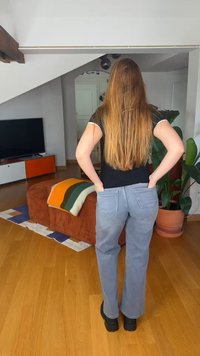 Sieviete ar gariem brūniem matiem stāv ar rokām pelēku džinsu aizmugurējās kabatās mūsdienīgā viesistabā ar augiem un televizoru.