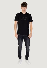 Maglietta nera in cotone con logo EA7 testurizzato sul davanti, abbinata a jeans scuri e sneakers bianche, per un look casual.