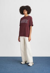 Bordo Nike T-krekls ar īsām piedurknēm un lielu logo, apvienots ar krēma platām biksēm un baltām kedām uz zila paklāja fona.