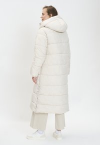 Manteau long rembourré en beige clair avec une capuche, présentant un design matelassé et des boutons sur les côtés. Associé à un pantalon beige clair et des baskets.
