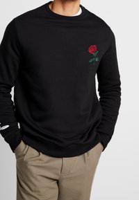 Felpa nera in misto cotone, con un piccolo ricamo di rosa rossa sul lato sinistro del petto e polsi e orlo a coste.