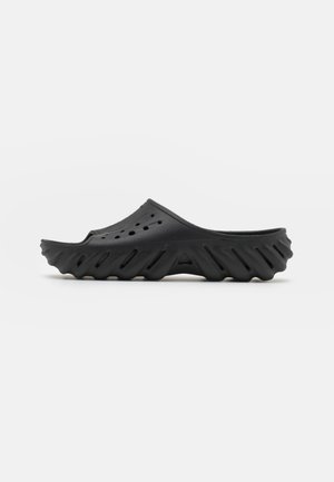 Black slip-on clog made of rubber, featuring a textured sole with a wavy design, ventilation holes on the upper, and a low, rounded profile.