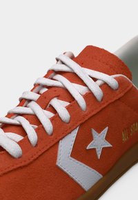 Κοντινό πλάνο πορτοκαλί αθλητικού παπουτσιού από σουέτ με λευκά κορδόνια, λευκό αστέρι και λογότυπο σε σχήμα βέλους, και καφέ σόλα.