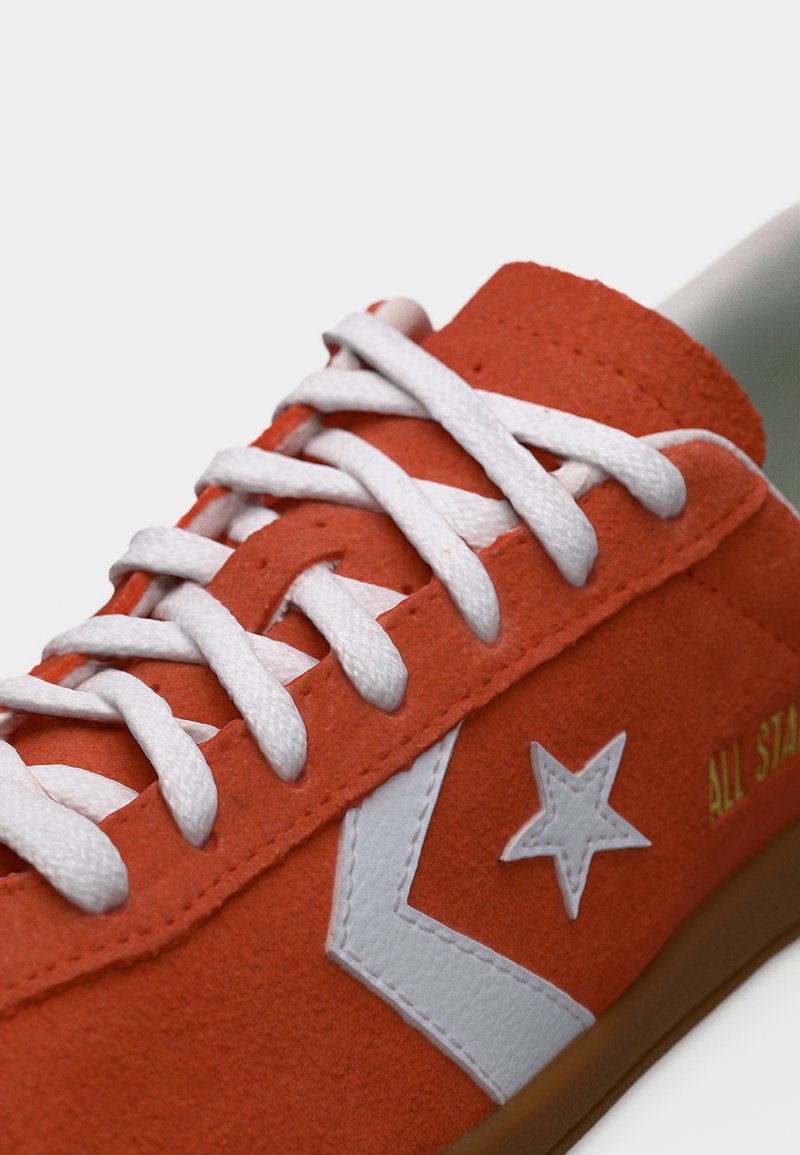 Primer plano de zapatilla de ante naranja con cordones blancos, estrella blanca y logo de chevrón, y suela marrón.