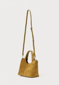 Piccola borsa in suede color sabbia con manici corti e tracolla lunga regolabile appesa su uno sfondo bianco semplice.