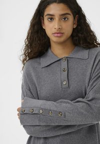 Young woman with curly hair wearing a gray long-sleeve sweater featuring buttons on the collar and sleeve cuffs, arms crossed.