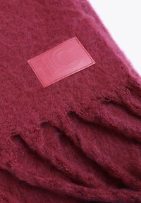 Burgundska vuna šal s resicama i pravokutnom crvenom kožnom oznakom s utisnutim inicijalima. Mekana tekstura i topli izgled.