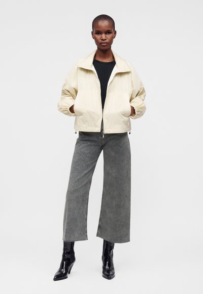 Femme portant une veste crème zippée, un top noir, un jean gris à jambes larges et des bottines noires à talons, debout les mains dans les poches.