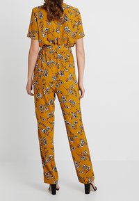 Květinový overal v hořčičné žluté barvě, s krátkými rukávy, elastickým pasem a černým květinovým potiskem. Měkký materiál s rovnými nohavicemi.