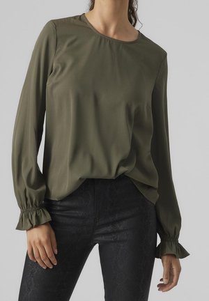 Vrouw die een olijfgroene blouse met lange mouwen en geplooide ruches bij de manchetten draagt, gecombineerd met zwarte textuurbroeken, staand tegen een effen grijze achtergrond.