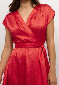 Röd och rosa mönstrad omlotklänning med korta ärmar, knuten i midjan, bärs av en person som står framför en enfärgad bakgrund.