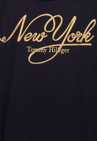 Marineblaue Baumwoll-T-Shirt, das in goldener Kursivschrift "New York" und darunter in Standardfont "Tommy Hilfiger" zeigt.