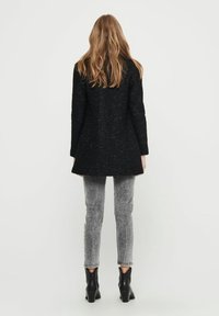 Schwarzer Mantel mit strukturierter Oberfläche und geradem Schnitt. Kombiniert mit grauen Jeans und schwarzen Stiefeletten mit Absatz.