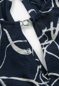 Tissu bleu marine avec une finition texturée, arborant un motif abstrait blanc. Un détail de fermeture avec un anneau en métal est visible près de la couture du col.