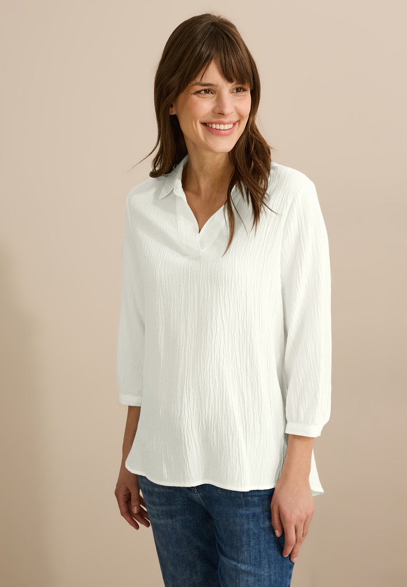 Camisa blanca de tejido texturizado, con cuello y mangas tres cuartos, combinada con jeans azules.