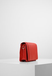 Červená kožená kabelka se strukturovaným tvarem, s klopovým uzavíráním a kovovými akcenty. Hladký povrch s minimálními designovými detaily.
