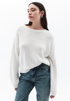 White knitted sweater with a wide neckline and ribbed cuffs, paired with blue denim jeans. Soft texture and relaxed fit.