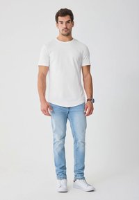 Vit t-shirt med korta ärmar i lätt bomullsmaterial, bärs med ljusblå jeans och vita sneakers, står mot en enfärgad bakgrund.