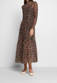 Schwarzes Blumenkleid im Maxistil mit langen transparenten Ärmeln, hohem Ausschnitt und gerafftem Taillenbereich. Mit pinkfarbenem Blumendruck und gestuftem Saum. Getragen mit cremefarbenen Schuhen.