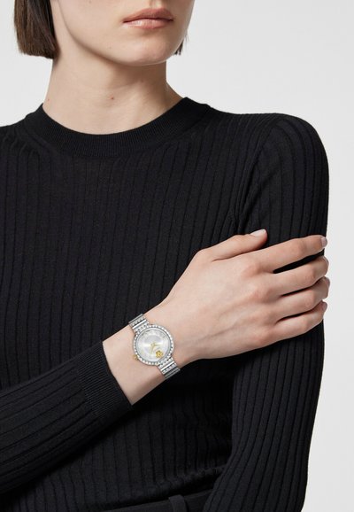 Sudraba pulkstenis ar apaļu baltu seju, zeltainiem akcentiem un rāmīti, kas rotāts ar rhinestones; uz rokas, kas valkā melnu ribotu džemperi.