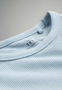 Lyseblå bomuld T-shirt med ribbet tekstur, med rund halsudskæring, syede kanter og en etiket, der lyder "SET MADE IN BANGLADESH."