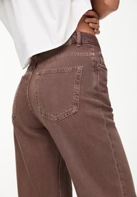 Jeans en denim marron avec une taille haute, dotés de deux poches arrière et d'une finition texturée, partiellement visibles sous un haut blanc.