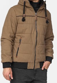 Braune gesteppte Jacke mit Kapuze, schwarzem Reißverschluss und geflochtenen Kordeln. Verfügt über gerippte Bündchen und zwei seitliche Reißverschlusstaschen.
