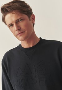 Czarna bluza z teksturowanymi, wypukłymi literami tworzącymi napis "Miś Uszatek." Wzór zawiera okrągły dekolt i ściągacze na mankietach.