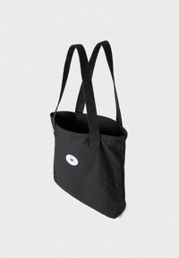 Tote bag en toile noire avec deux longues anses, présentant un logo circulaire avec une étoile bleue et une bordure blanche sur un côté. Texture douce.