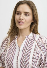 Biely blúz s viacfarebnými geometrickými vzormi, zapínaním na gombík a naťahovacími rukávmi. Textúrovaná tkanina s precíznym detailom po celej ploche.