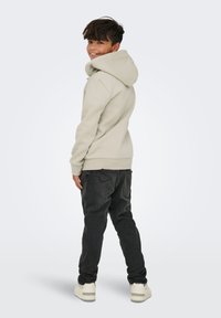 Un garçon souriant par-dessus son épaule, portant un hoodie beige clair, un jean noir et des baskets blanches, se tenant devant un fond clair.