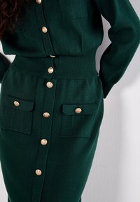 Robe en tricot verte avec une taille cintrée, deux poches avant et des boutons en or. Présente une texture côtelée et des manches longues.