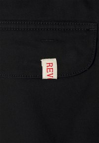 Zwarte stoffen achtergrond met een wit label waarop in rode tekst "REV" is gestikt in een zak. De stiksels zijn zichtbaar langs de randen.