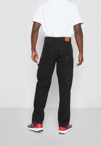 Czarne spodnie cargo z wieloma kieszeniami, kontrastowym przeszyciem i skórzaną naszywką na plecach. Noszone z białym t-shirtem i tenisówkami.