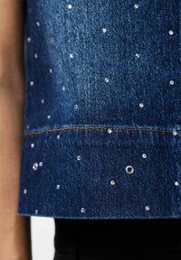 Jeansväst i en mörkblå tvätt, prydd med klara rhinestones och silverdetaljer, med rak fåll och synlig sömnad.