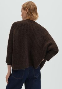 Donna con capelli biondi corti che indossa un maglione largo di colore marrone scuro con trama e jeans blu scuro, vista da dietro su uno sfondo neutro.