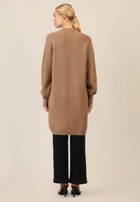 Maglione cardigan lungo marrone a coste con spalle ribassate, indossato sopra pantaloni neri a gamba larga. Tessuto liscio e vestibilità comoda, design fino al ginocchio.