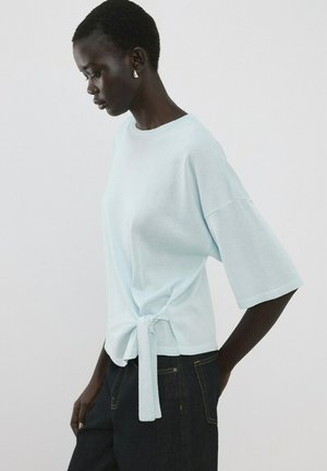 Mujer con camiseta de manga corta color azul claro con detalle de lazo lateral y jeans oscuros, de pie de lado contra un fondo blanco liso.