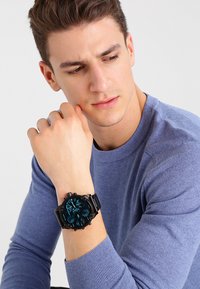 Černé náramkové hodinky s kulatým modr�ým ciferníkem, zobrazujícími důmyslné detaily, nošené na zápěstí, na jednotném pozadí, s modrým svetr.