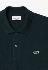 Tumma vihreä polo-paita, jossa on teksturoitu kangas, kolme napin läpivientiä ja pieni valkoinen brodeerattu Lacoste-logo vasemmassa rinnassa.