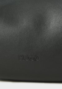 Černá kožená taška s hladkým povrchem, na přední straně s embosovaným logem "HUGO". Jednoduchý, cylindrický design bez dalších kovových prvků.