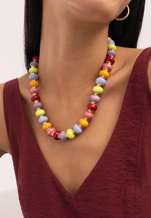 Collar con grandes cuentas redondas y coloridas en rojo, naranja, amarillo, verde, azul, púrpura y dorado, llevado con una camiseta sin mangas de color granate y un pendiente de aro dorado.