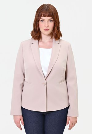 Donna con capelli rossi fino alle spalle, che indossa un blazer beige sopra una maglietta bianca e jeans blu scuro, in piedi davanti a uno sfondo bianco.