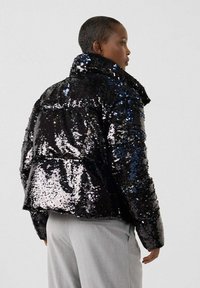 Personne de profil portant une doudoune noire à sequins brillants et un pantalon gris clair sur un fond uni clair.