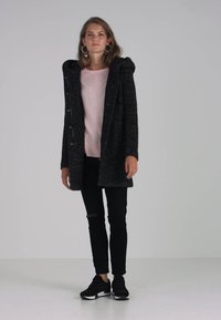 Črn volnen plašč s kapuco, gumbasto zapiranje spredaj in strukturirano tkanino; v kombinaciji z roza puloverjem, črnimi raztrganimi jeans hlačami in črnimi teniskami.