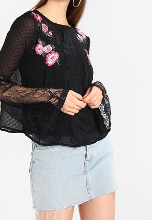 Vrouw draagt een zwarte doorschijnende kanten blouse met roze rozenborduursel, gecombineerd met een lichtblauwe denim minirok.