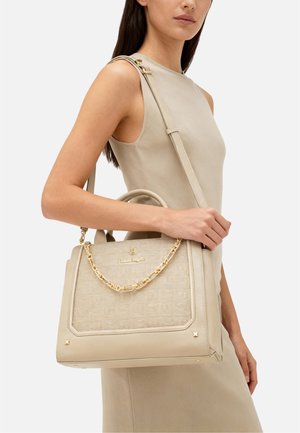 Donna con vestito senza maniche beige che tiene una borsa beige con dettagli in catena d'oro sulla spalla.