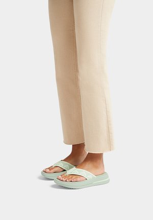 Lichtgroene flip-flops met gestructureerde rubberen zolen en brede banden, gecombineerd met beige cropped broeken. De banden hebben een subtiel patroon.