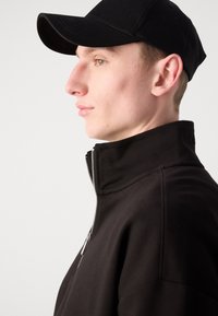 Crna jakna na patentni zatvarač s visokim ovratnikom, izrađena od glatkog materijala. Nosena s crnom kapom, fokusira se na bočni profil.