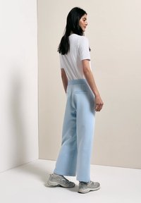Femme aux longs cheveux foncés, portant un haut blanc à manches courtes, un pantalon large bleu clair et des baskets argentées, debout contre un mur beige uni.
